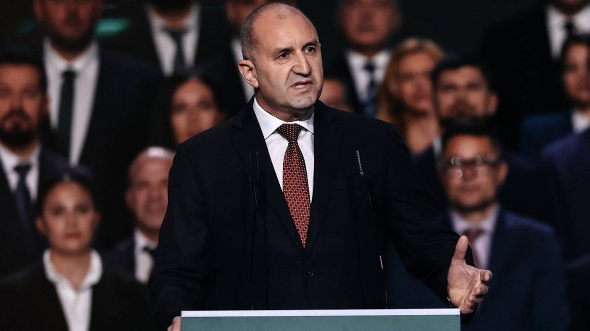 Polityk w garniturze przemawia za m&oacute;wnicą, otoczony przez innych ludzi.