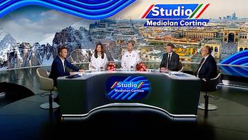 Cztery osoby siedzą przy stole w studiu telewizyjnym, w tle widok na miasto i góry, nad stołem logo "Studio Mediolan Cortina".