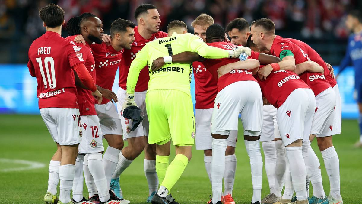 Grupa piłkarzy w czerwonych strojach otacza jednego zawodnika w żółtym stroju bramkarskim, ściskając się i wspierając w geście jedności.