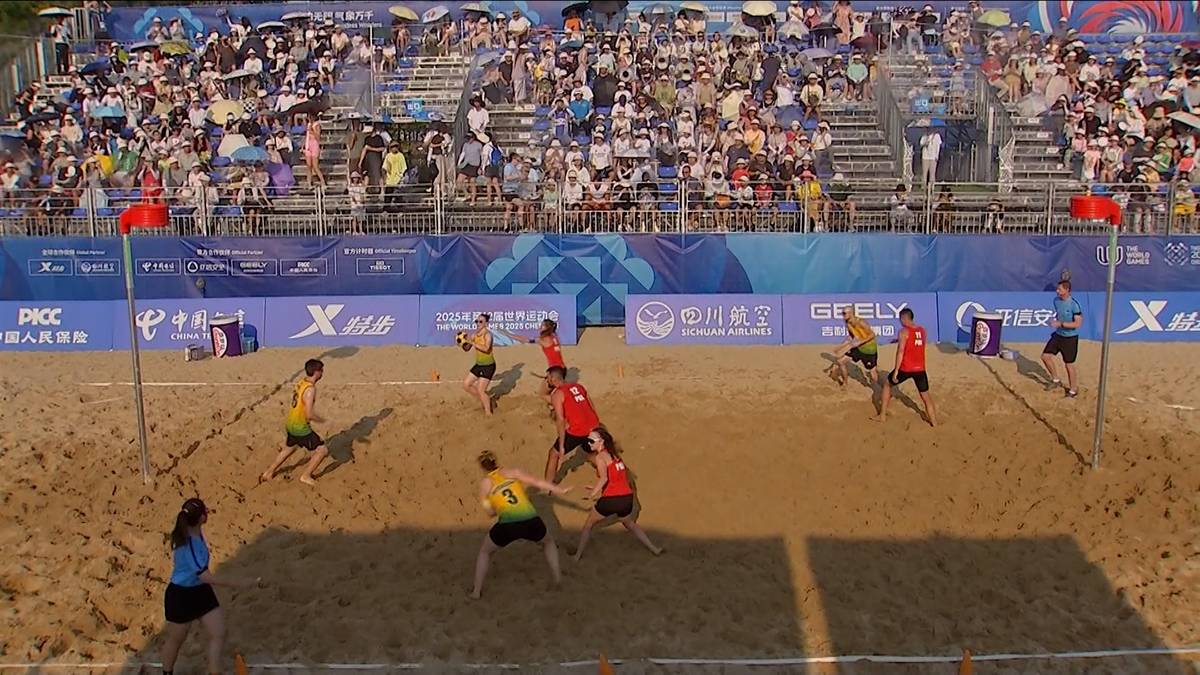 Gracze w korfball plażowy rozgrywają mecz na piaszczystym boisku. W tle znajdują się trybuny wypełnione widzami.