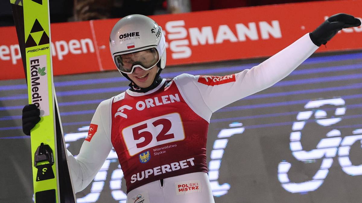 Skoczek narciarski w białym kasku i białej kurtce z czerwonymi elementami, trzymający nartę.