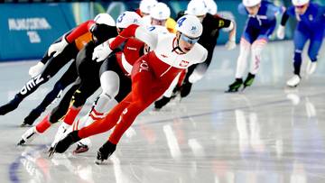 Wyścig łyżwiarstwa szybkiego, zawodnik w biało-czerwonym stroju z polską flagą na prowadzeniu.