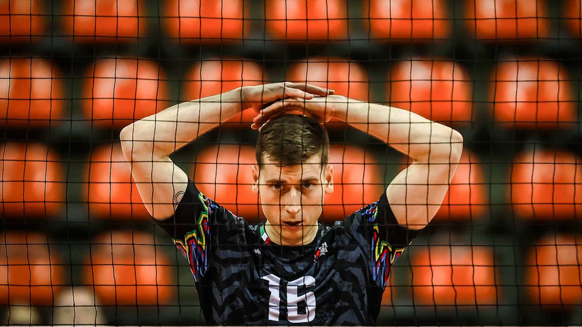 Młody mężczyzna w czarnej koszulce z numerem 16 na plecach, z rękami splecionymi na głowie, stoi na tle rzędu pomarańczowych krzesełek.