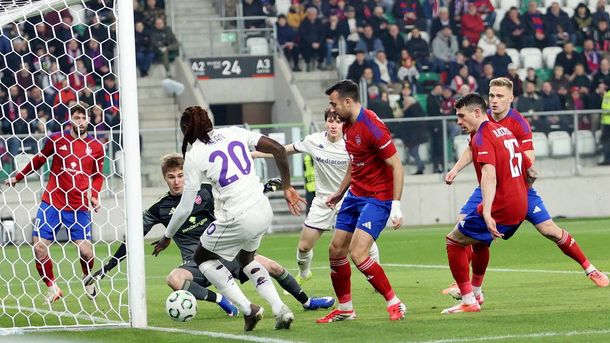 Piłkarze w akcji podczas meczu piłki nożnej, jeden z zawodnik&oacute;w w białej koszulce z numerem 20 pr&oacute;buje zdobyć bramkę, w tle inni zawodnicy i trybuny stadionu.