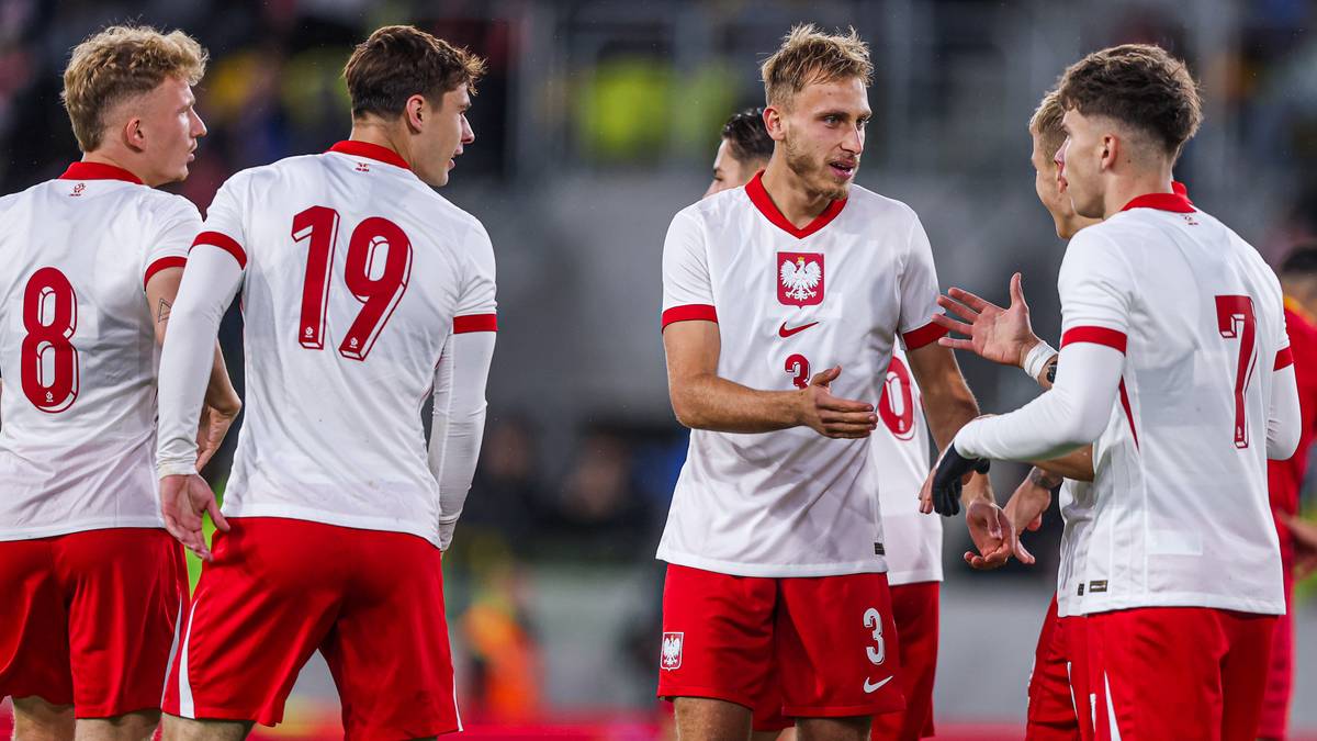 Pięciu zawodników reprezentacji Polski w piłce nożnej w białych koszulkach z czerwonymi detalami i numerami na plecach, rozmawiających ze sobą podczas meczu.