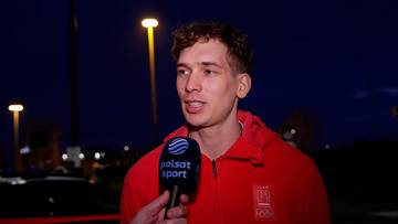 Młody mężczyzna w czerwonej bluzie z kapturem udziela wywiadu, trzymając mikrofon z napisem "Polsat Sport". W tle widoczne są dwie latarnie uliczne oświetlające nocną scenerię.