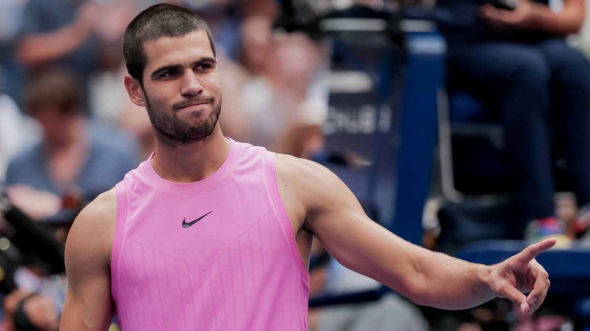 Carlos Alcaraz w różowej koszulce polo, ze wzrokiem skierowanym w bok i lekko podniesioną brwią, pokazuje palcem w prawo.