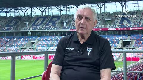 Portret starszego mężczyzny w czarnej koszulce z logo klubu sportowego, siedzącego na tle stadionu piłkarskiego z widownią.