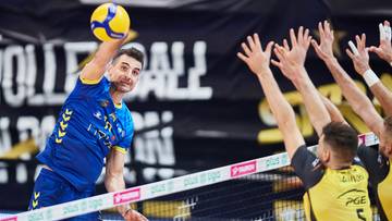 Siatkarz w niebieskiej koszulce z ż&oacute;łtymi akcentami wykonuje zagrywkę, podczas gdy przeciwnicy w ż&oacute;łtych strojach pr&oacute;bują go zablokować.