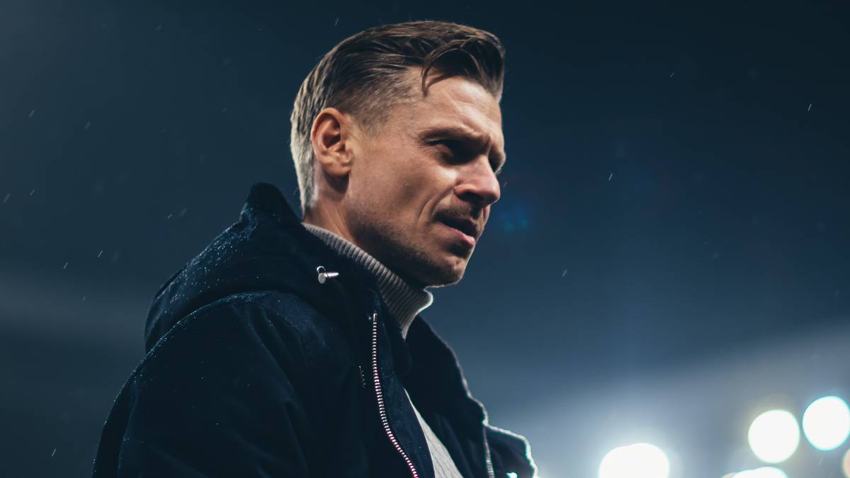 Mężczyzna w kurtce i golfie na tle oświetlonego stadionu.