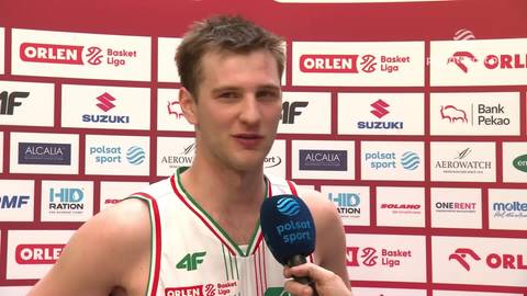 Mężczyzna w białej koszulce z zielonymi i czerwonymi paskami, trzymający mikrofon, udziela wywiadu.