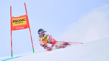 Narciarka w białym i r&oacute;żowym kombinezonie zjazdowym, kasku i goglach, z nartami na nogach, przejeżdża przez trasę narciarską obok czerwonej bramki z ż&oacute;łtym znakiem "ZGONC"