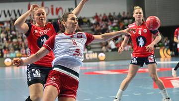 Trzy zawodniczki piłki ręcznej w akcji. Jedna z nich w biało-czerwonym stroju pr&oacute;buje rzucić piłkę, podczas gdy dwie przeciwniczki w czerwonych strojach pr&oacute;bują ją zablokować.