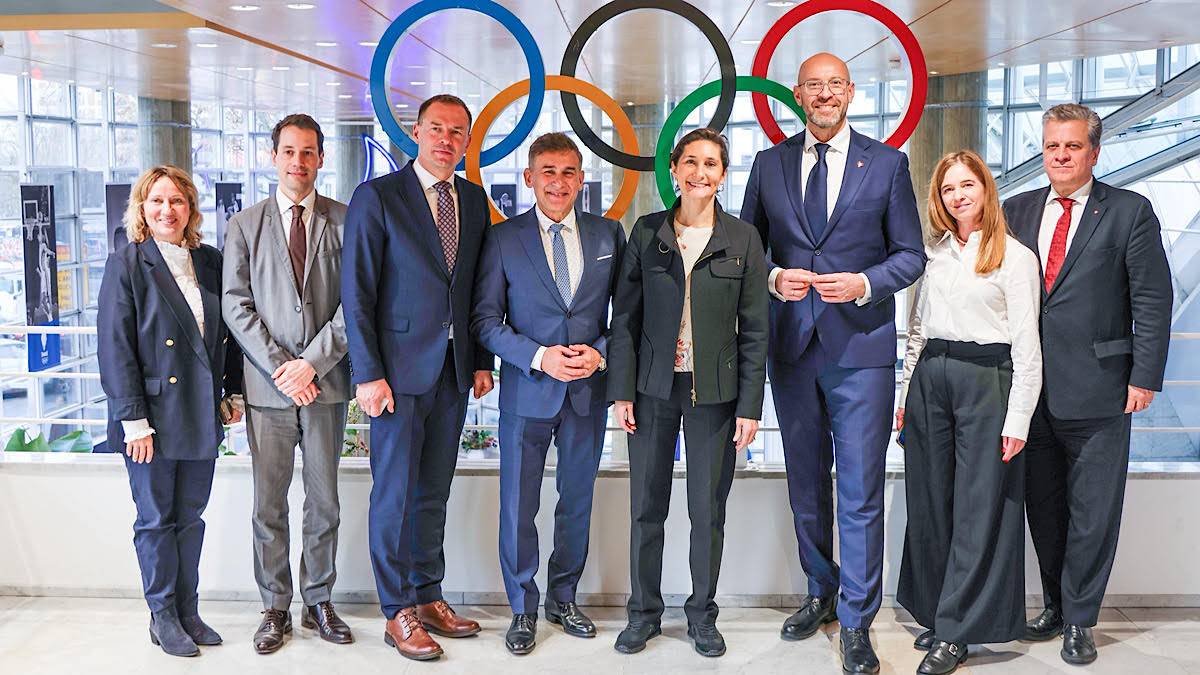 Grupa osób w strojach formalnych stoi przed logo igrzysk olimpijskich.