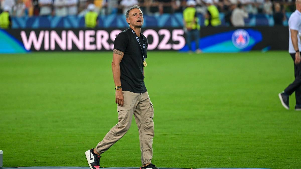 Mężczyzna w średnim wieku, ubrany w czarną koszulkę polo, beżowe spodnie typu cargo i biało-czarne buty sportowe, idzie po zielonej murawie stadionu piłkarskiego. Ma na szyi złoty medal. W tle widoczny jest ekran z napisem "WINNERS 2025".