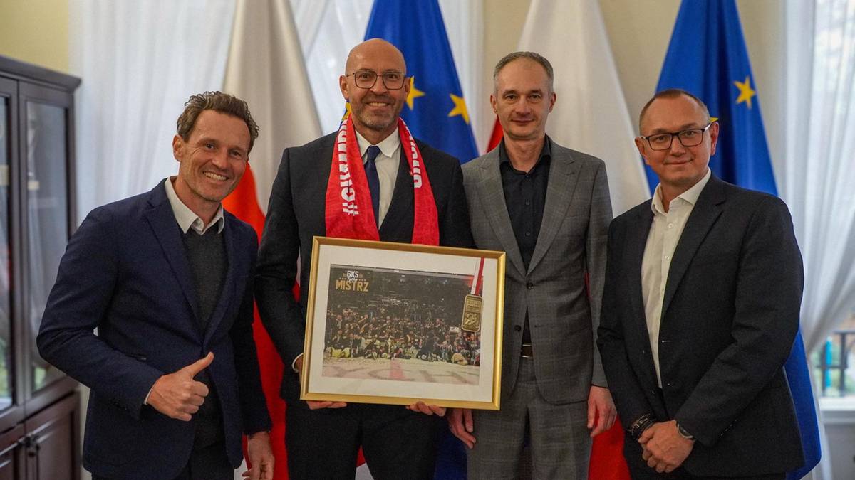 Czterech mężczyzn stoi przed flagami, trzymając w ramce zdjęcie przedstawiające grupę osób. Mężczyźni są ubrani w garnitury i koszule. Jeden z nich ma na sobie czerwony szalik.