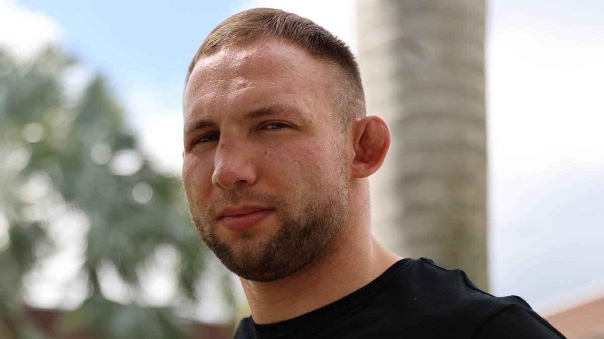 Mężczyzna w czarnej koszulce z napisem "PITBULL" na pomarańczowym tle, z zaciśniętymi pięściami.