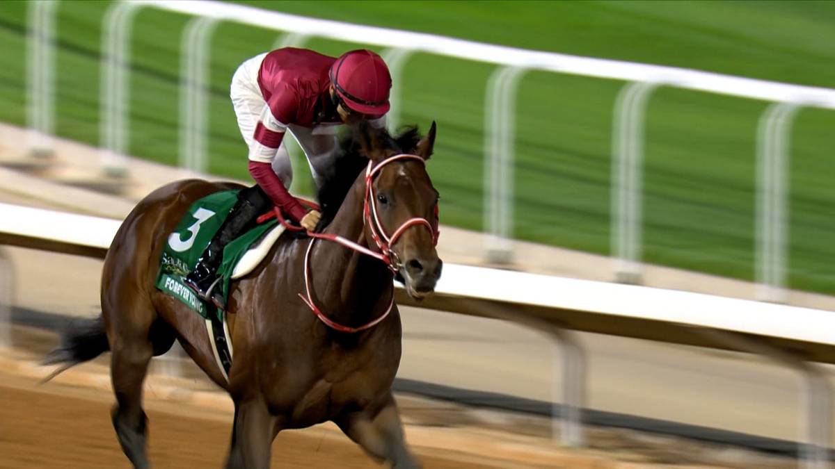 Koń wyścigowy z dżokejem na grzbiecie biegnie po piaszczystym torze, z zielonym tłem i bandami toru.