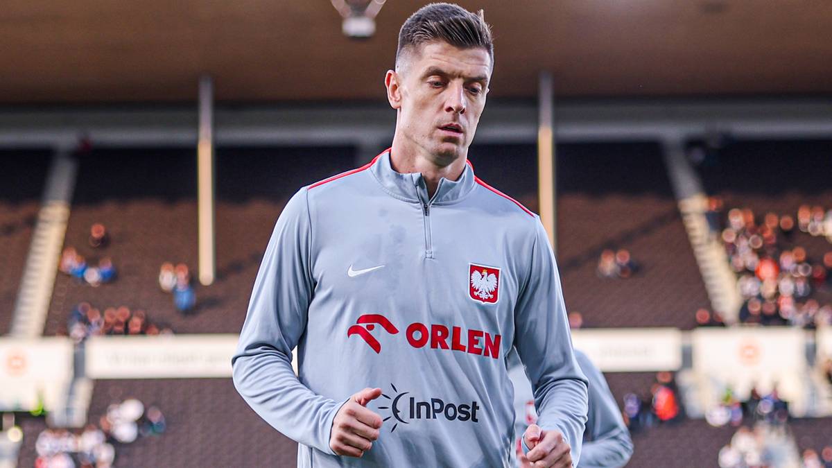 Piłkarz Krzysztof Piątek w szarym dresie reprezentacji Polski z logo Orlen i InPost, stojący na stadionie.