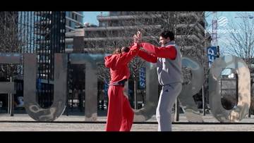 Dwoje młodych ludzi w strojach sportowych, kobieta w czerwonym dresie i mężczyzna w szarym dresie, tańczy przed dużymi metalowymi literami "EUROPA".