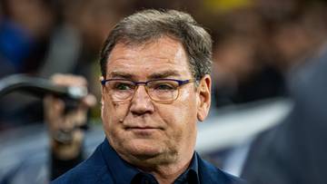 Trener piłkarski w okularach, ubrany w ciemny garnitur, w skupieniu obserwuje wydarzenia na boisku.