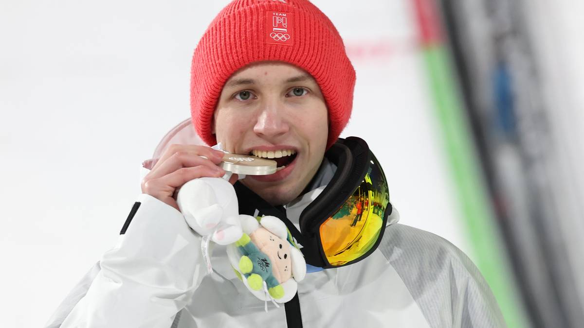 Portret mężczyzny w czerwonej czapce i goglach narciarskich, trzymającego medal w ustach.