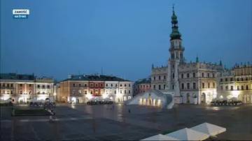 Rynek w Zamościu o poranku, widoczne zabytkowe kamienice, ratusz i wieża. Plac jest pusty, z mokrą nawierzchnią i kilkoma białymi parasolami na pierwszym planie.