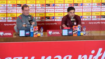 Dwaj mężczyźni siedzą za stołem podczas konferencji prasowej reprezentacji Polski. Na stole znajdują się mikrofony, butelki z wodą i materiały promocyjne. W tle widnieje ściana z logo sponsorów i napisem "EUROPEAN QUALIFIERS".