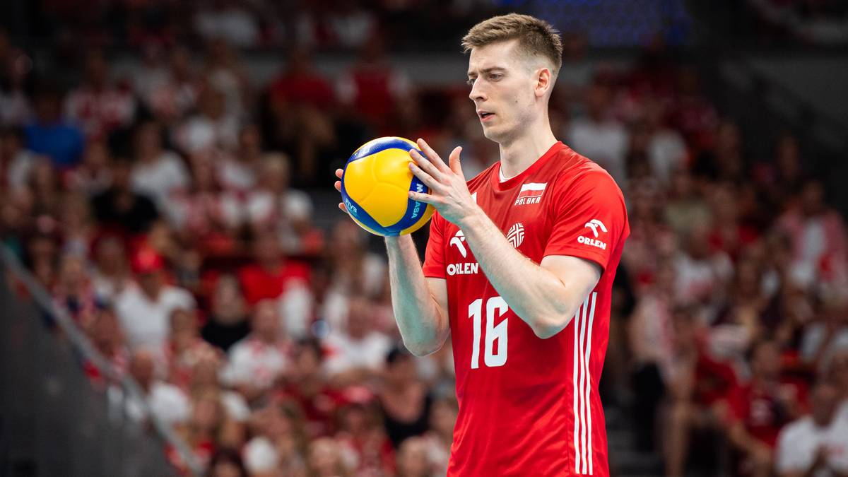 Młody siatkarz w czerwonej koszulce reprezentacji Polski z numerem 16 trzyma piłkę do siatkówki w obu rękach, patrząc przed siebie z niezadowoloną miną. W tle widać trybuny wypełnione kibicami.