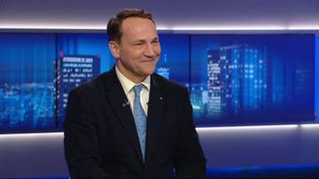 Radosław Sikorski w garniturze i krawacie, uśmiechający się w studiu telewizyjnym z nocnym krajobrazem miasta w tle.