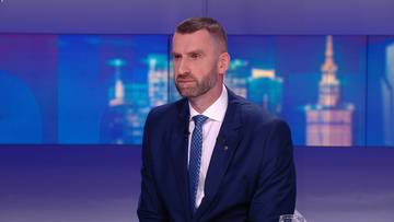 Mężczyzna w ciemnym garniturze i niebieskim krawacie siedzi, patrząc na wprost. W tle znajduje się niebieski ekran z widokiem na miasto.