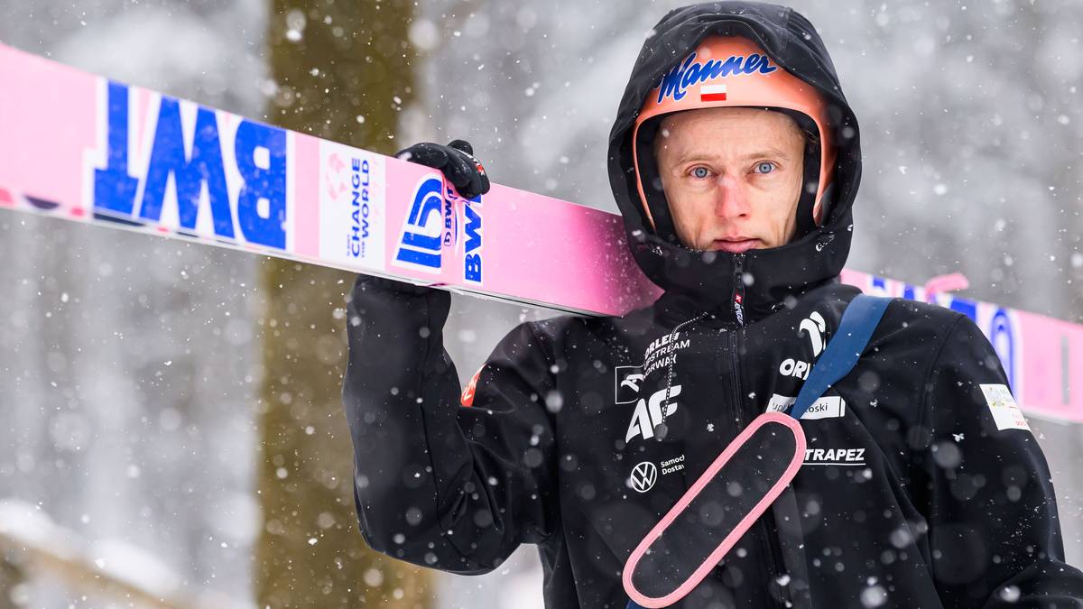 Skoczek narciarski w kasku i czarnej kurtce trzyma narty na ramieniu, na nartach widoczny napis BWT.