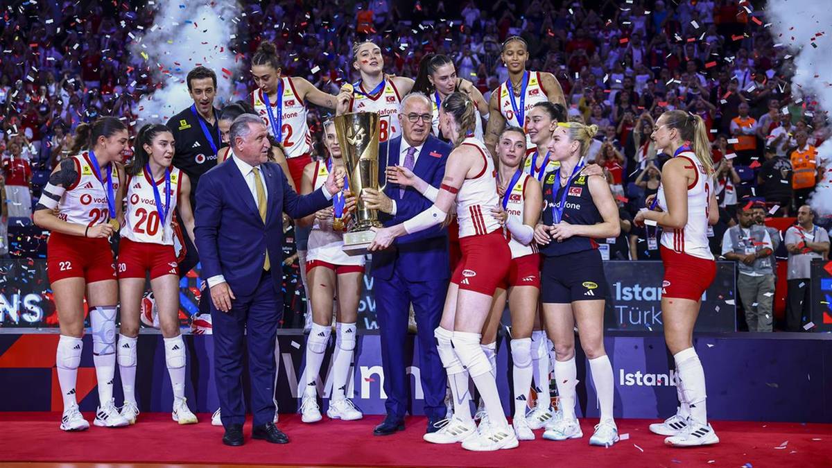 Drużyna siatkarek świętuje zwycięstwo, unosząc w górę puchar. Na zdjęciu widoczni są zawodnicy w strojach sportowych, trener oraz część publiczności na trybunach.