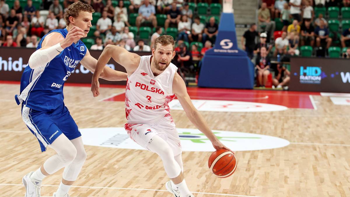 Dwóch koszykarzy grających mecz na boisku halowym. Jeden z nich w białej koszulce z napisem 'POLSKA' prowadzi piłkę prawą ręką, drugi w niebieskiej koszulce próbuje go zatrzymać.