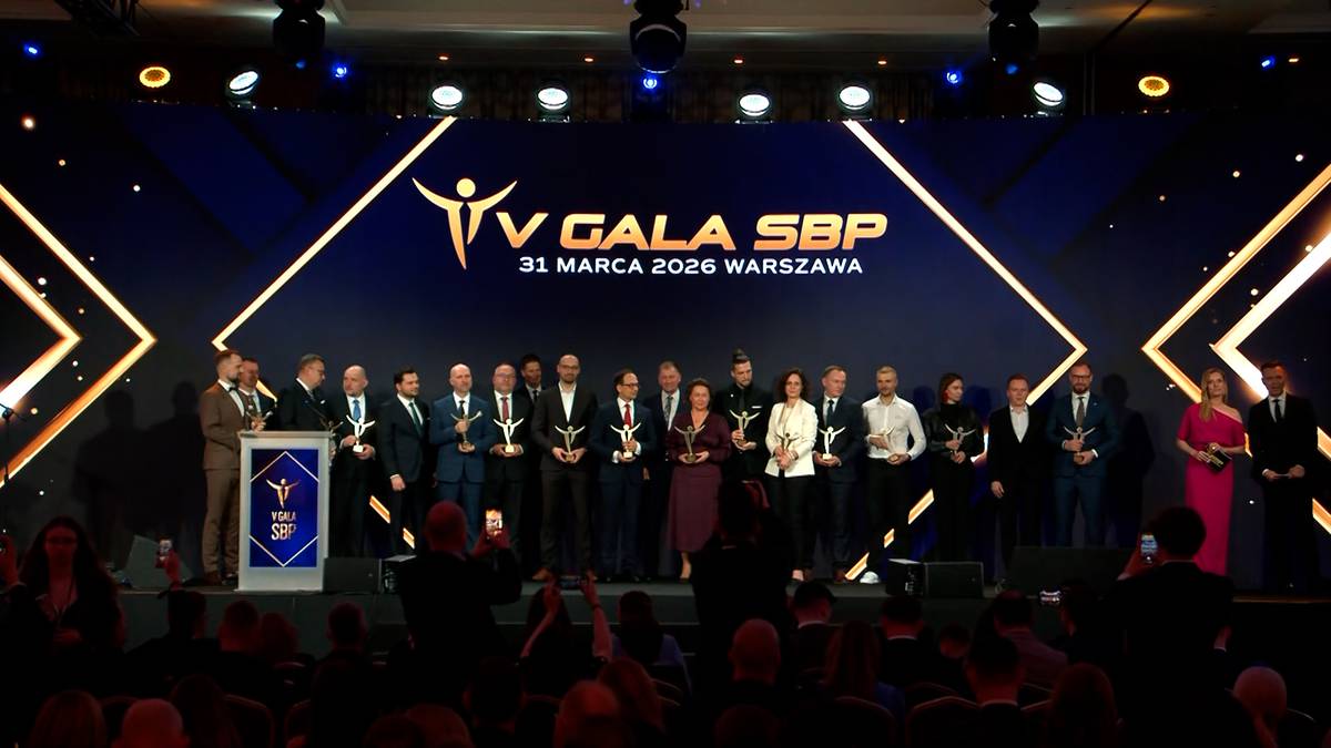Grupa os&oacute;b stoi na scenie podczas gali, na kt&oacute;rej wręczane są nagrody. W tle widnieje duże logo "V GALA SBP".