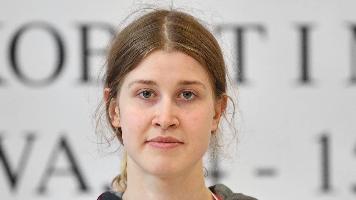 Portret młodej kobiety z jasnymi włosami, patrzącej prosto przed siebie. Tło jest niewyraźne, z literami i cyframi.