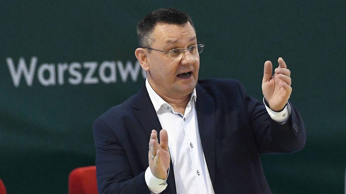 Trener koszykówki w garniturze i białej koszuli gestykuluje rękami.