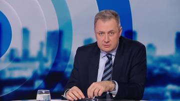 Mężczyzna w garniturze siedzi przy stole, przed nim znajduje się szklanka wody.