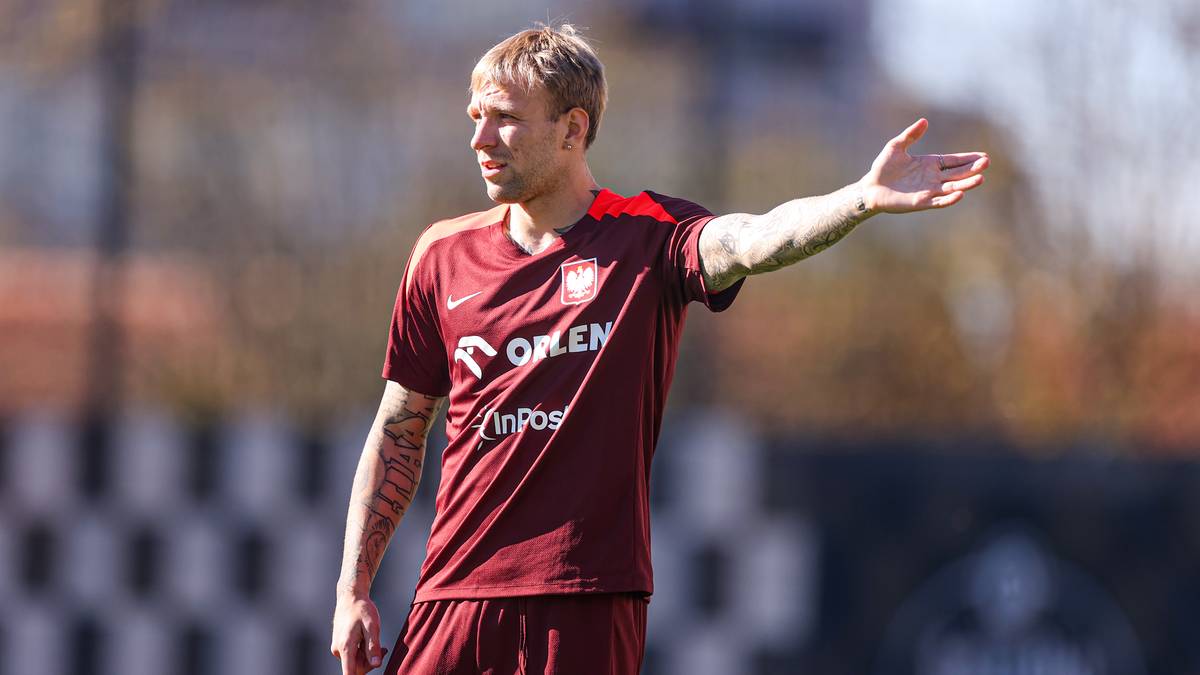 Piłkarz w czerwonej koszulce z białym logo i napisami wykonuje gest prawą ręką.