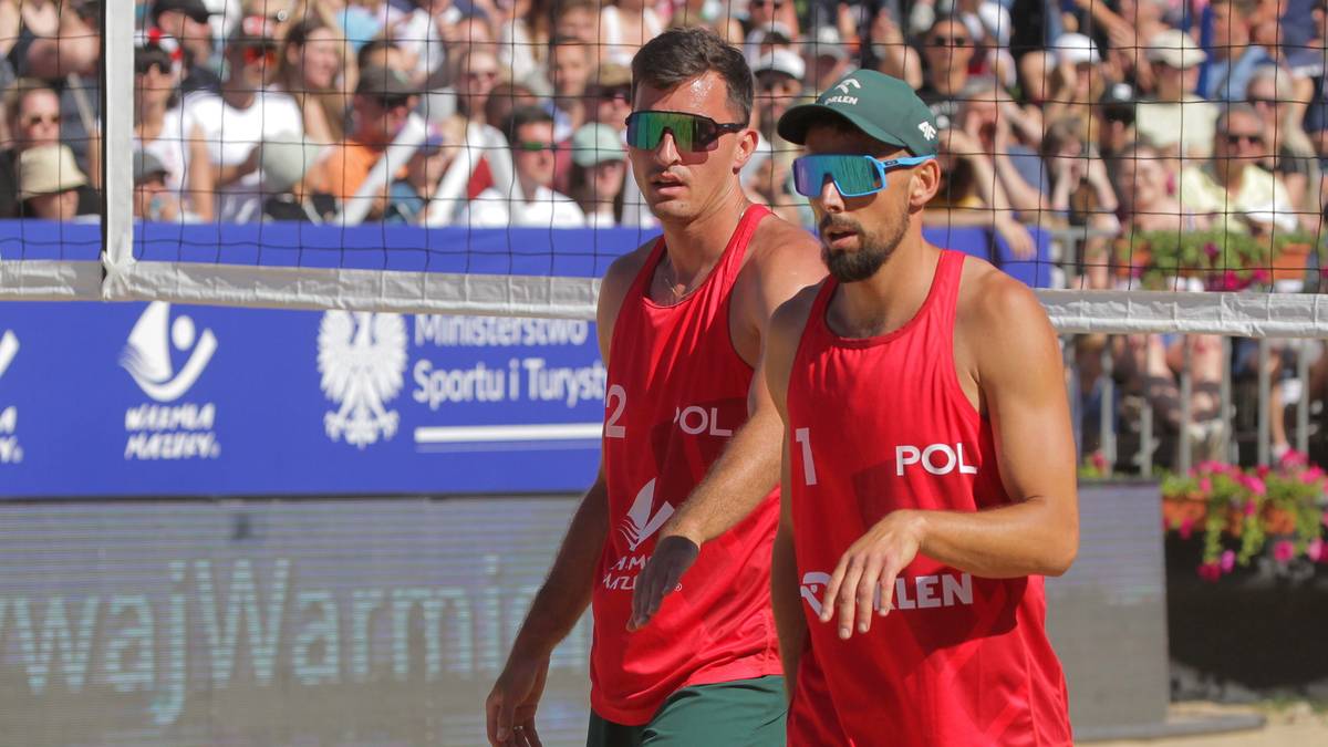Dwóch polskich siatkarzy plażowych w czerwonych koszulkach idzie po piasku, z trybunami kibiców w tle.