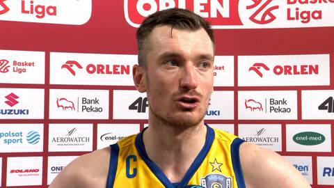 Portret Daniela Gołębiowskiego, koszykarza, w żółtej koszulce z niebieskimi elementami, na tle ściany z logotypami sponsorów.