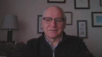 Portret mężczyzny w okularach i ciemnej kurtce, siedzącego przed ścianą z obrazkami i lampą.