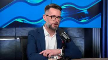 Mężczyzna w okularach i granatowej marynarce siedzi przy stole, m&oacute;wiąc do mikrofonu.