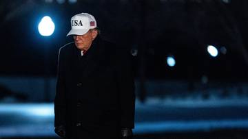Donald Trump w czapce z napisem USA, ubrany w czarny płaszcz, stoi na zewnątrz w nocy.