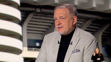 Portret mężczyzny w średnim wieku, z siwymi włosami i zarostem, ubranego w jasnoszarą marynarkę, czarny golf i koszulę. Mężczyzna patrzy w bok i m&oacute;wi. W prawym dolnym rogu kadru widoczna jest kobieta prowadząca tłumaczenie na język migowy.