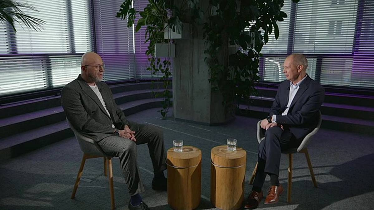 Dw&oacute;ch mężczyzn w garniturach siedzi naprzeciwko siebie na krzesłach w pomieszczeniu z przeszklonymi ścianami.