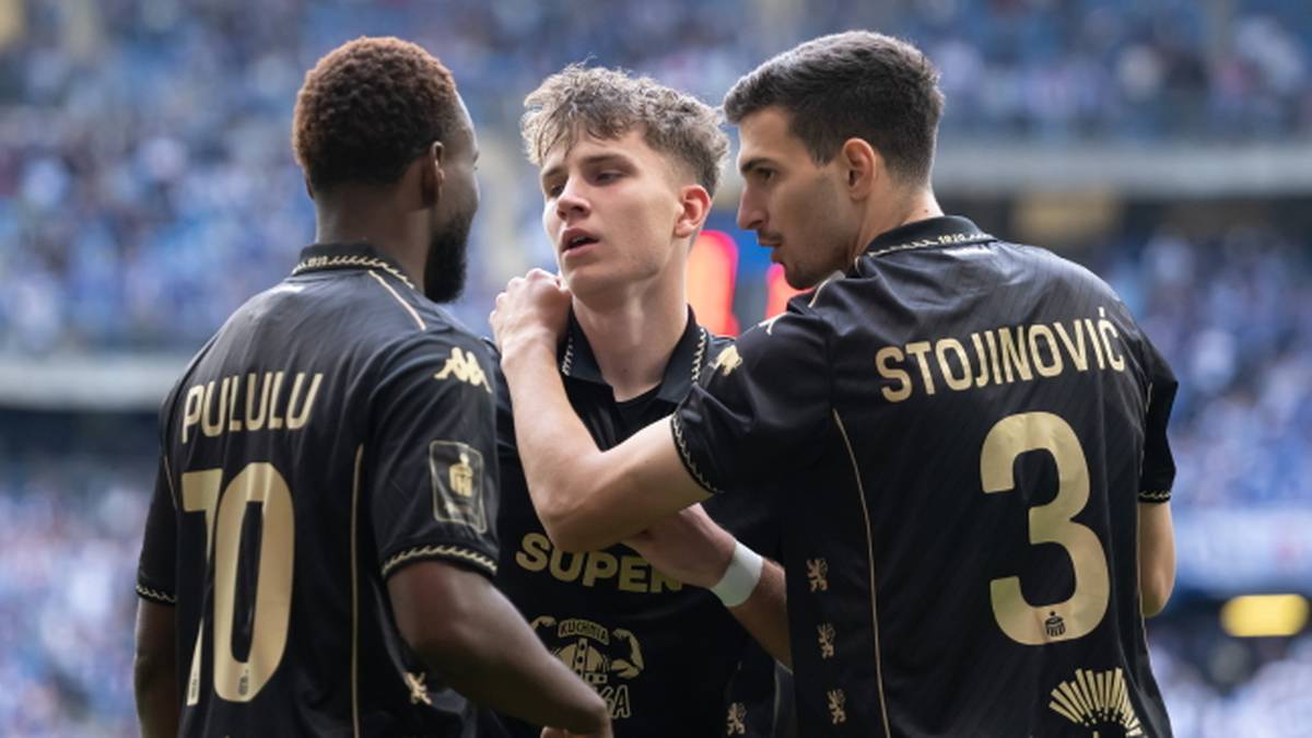 Trzech piłkarzy w czarnych koszulkach z żółtymi numerami i napisami dyskutuje na boisku.