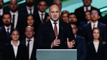 Mężczyzna w garniturze stoi za m&oacute;wnicą, przemawiając do grupy ludzi w tle.