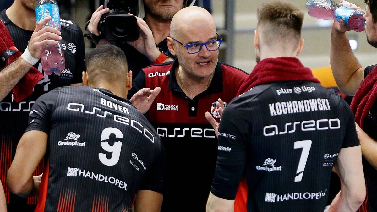 Trzech mężczyzn w strojach sportowych. Dw&oacute;ch w czarnych koszulkach z numerami 7 i 9, z tyłu głowy. Pomiędzy nimi mężczyzna w okularach, w czerwono-czarnej koszulce, trzymający ręce otwarte, wskazując nimi na rozmowę. Na koszulkach widoczne napisy: "ASSEC O" i "HANDLOPEX".