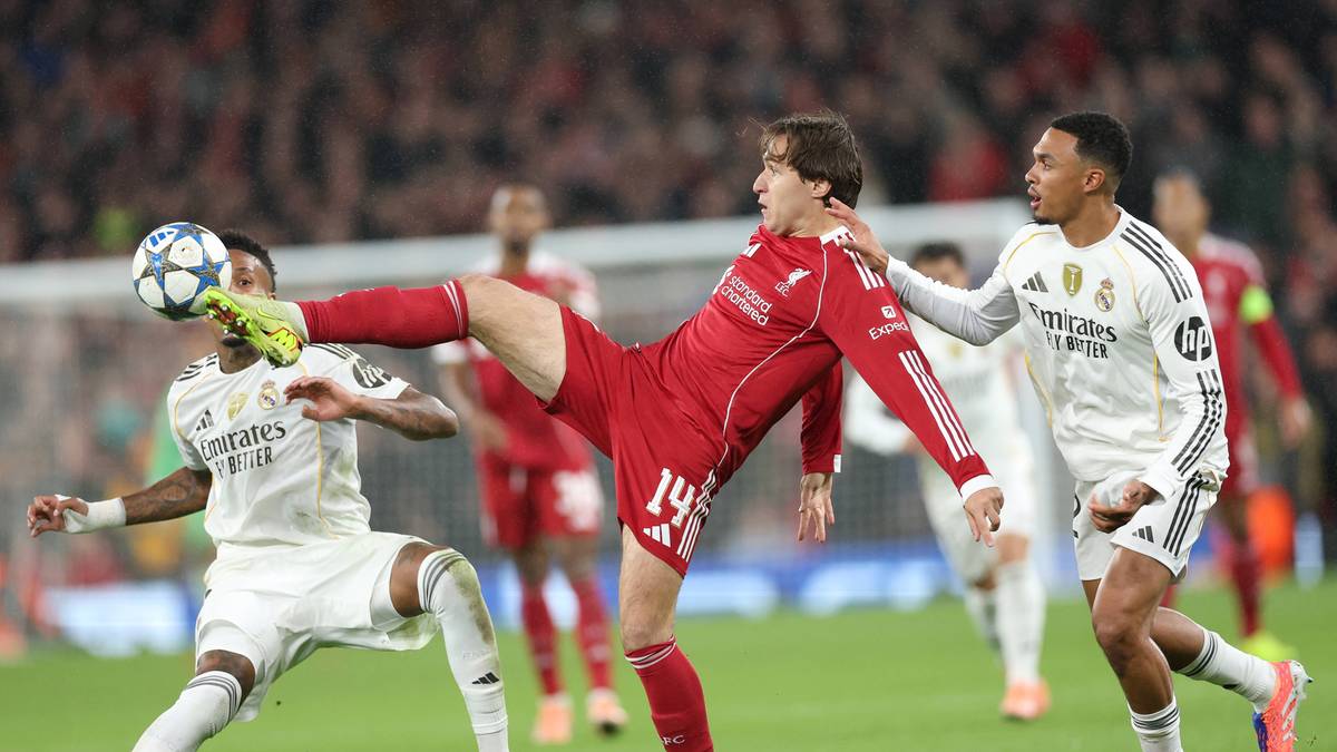 Trzech piłkarzy rywalizuje o piłkę na boisku, jeden w czerwonym stroju kopie piłkę, dwaj w białych strojach próbują go zatrzymać.